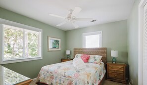 Un dormitorio con cama, dos mesitas de noche, ventilador de techo y dos ventanas con persianas.
