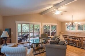 A spacious living room with a glass-top coffee table, a sofa, and a dining table with chairs. Large windows provide ample natural light and views of the outdoors.