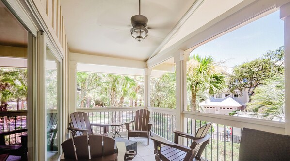 A covered outdoor area with a ceiling fan, a table, chairs, and a view of palm trees and a house.