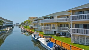 Un canal bordé de bateaux amarrés sur la rive droite, entouré d’immeubles à plusieurs étages dotés de balcons et d’une pelouse verdoyante.