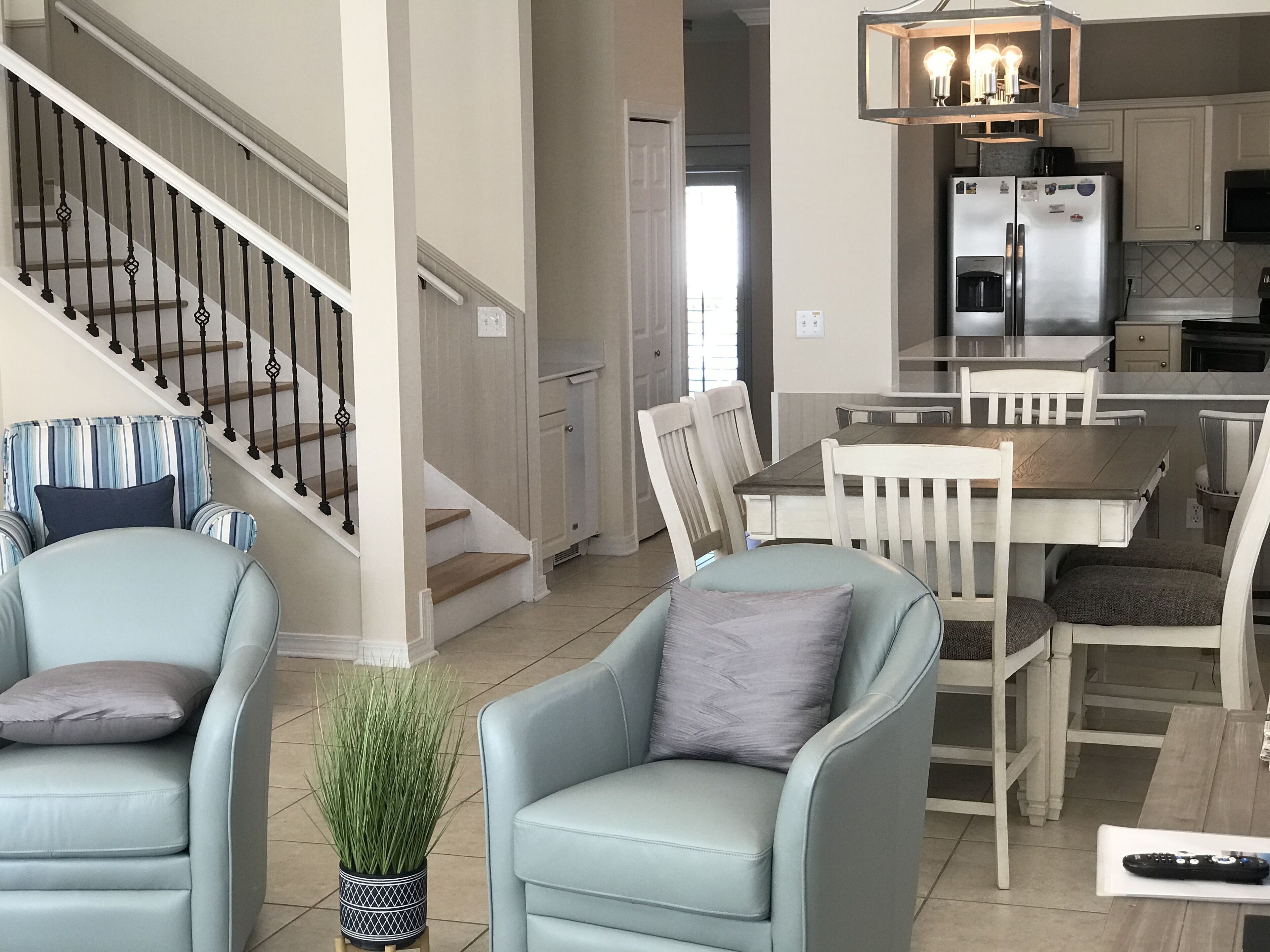A modern kitchen with a dining area, featuring a staircase, light blue chairs, a potted plant, and a remote control on the floor.