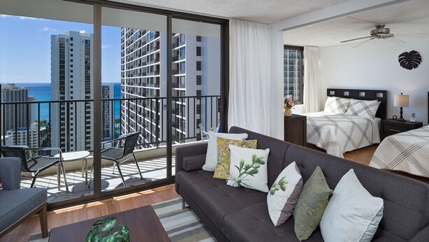 A modern hotel room with a balcony, a bed with a patterned bedspread, a sofa with decorative pillows, and a view of high-rise buildings and the ocean.