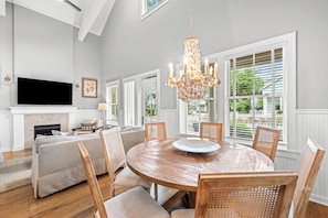A spacious living room with a fireplace, a large TV, a chandelier, and a dining area with a round table and chairs.