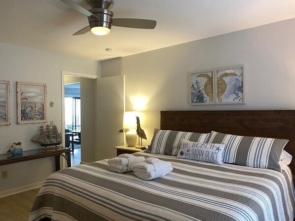 Ein ordentlich bezogenes Bett mit gestreifter Bettwäsche und einem Kissen mit Schriftzug, ein Deckenventilator und Kunstwerke an den Wänden.