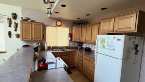 A kitchen with wooden cabinets, a white refrigerator, a clock on the wall, and a window with blinds. - Surfrider - Cozy Beach Cabin, Close to Beach, Pool Table (Pacific City)