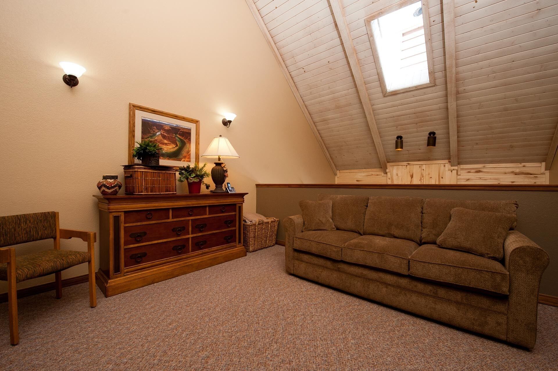 A cozy loft bedroom with a sloped ceiling, a wooden dresser, a brown sofa, and a small table with a lamp.