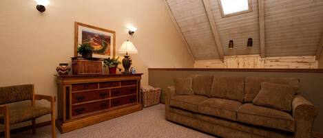 A cozy loft bedroom with a sloped ceiling, a wooden dresser, a brown sofa, and a small table with a lamp.