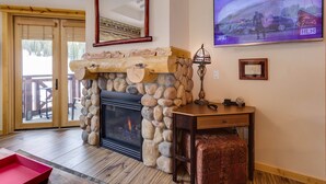 A room with a stone fireplace, a television mounted on the wall, and a wooden bench. - Steps to American Flyer lift & Stellar Mtn Views! Hot Tub, Free Wifi, Heated Pkg (Copper Mountain)