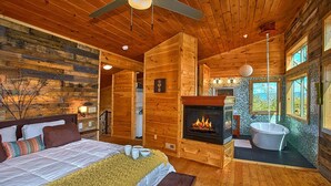 Ein Schlafzimmer in einer Blockhütte mit Kamin, einem großen Bett, einer Badewanne und Blick ins Freie.
