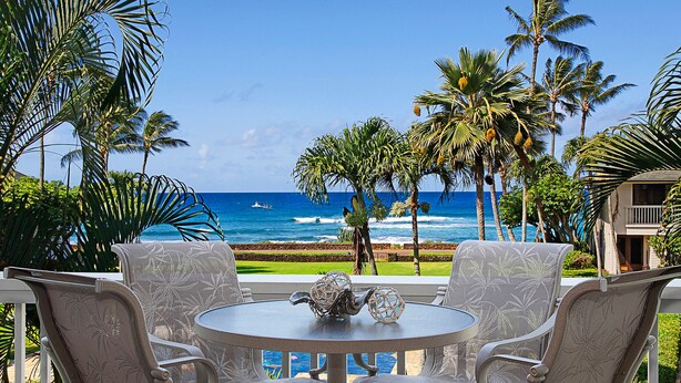 A patio with a round table and chairs, overlooking a beach with palm trees and a view of the ocean.