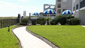 Un sendero serpentea por un césped cuidado, con sombrillas a rayas azules y blancas, una cerca y un edificio al fondo.