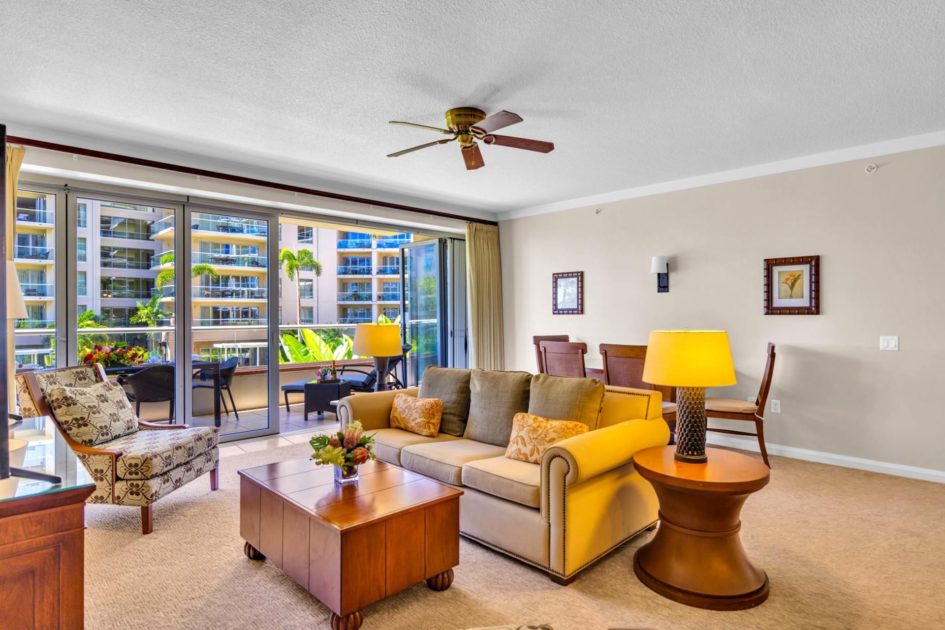 A spacious living room with a large sofa, a coffee table, and a dining area with chairs. There is a ceiling fan, a window with a view of a building, and a lamp on a side table.