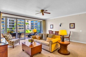 A spacious living room with a large sofa, a coffee table, and a dining area with chairs. There is a ceiling fan, a window with a view of a building, and a lamp on a side table.