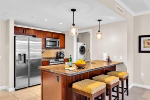 Une cuisine moderne dotée d’appareils électroménagers en acier inoxydable, d’un îlot central avec des tabourets de bar, d’un éclairage suspendu et d’un lave-linge en arrière-plan.