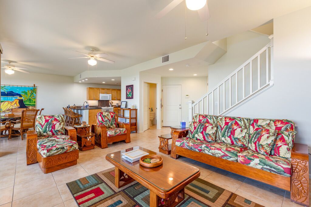 A spacious living room with a sofa, chairs, and a coffee table. There is a kitchen in the background with wooden cabinets and a microwave.
