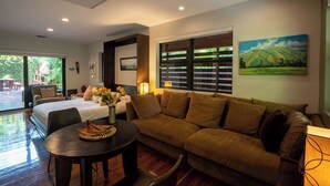 A modern living room with a sofa, a dining table, and a bed. There are large windows with blinds, a painting on the wall, and a view of greenery outside. - Coconut Grove Garden Cottage • Private Retreat with Free Parking & Full Kitchen (Miami)