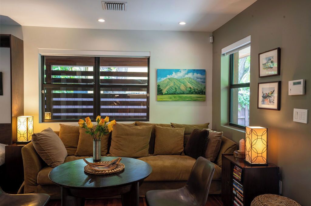 A cozy living room with a sofa, a round coffee table, and a bookshelf. There is a window with blinds, a painting on the wall, and a lamp on the side table.
