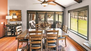 A dining area with a large wooden table, multiple chairs, a ceiling fan, and a view of a grassy landscape through large windows.