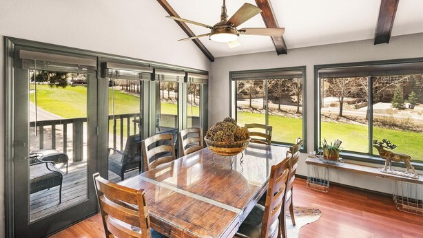 A dining area with a wooden table and chairs, a ceiling fan, and large windows offering a view of a grassy yard.