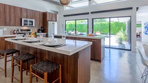 A modern kitchen with a large island, bar stools, and a view of the garden through large windows.