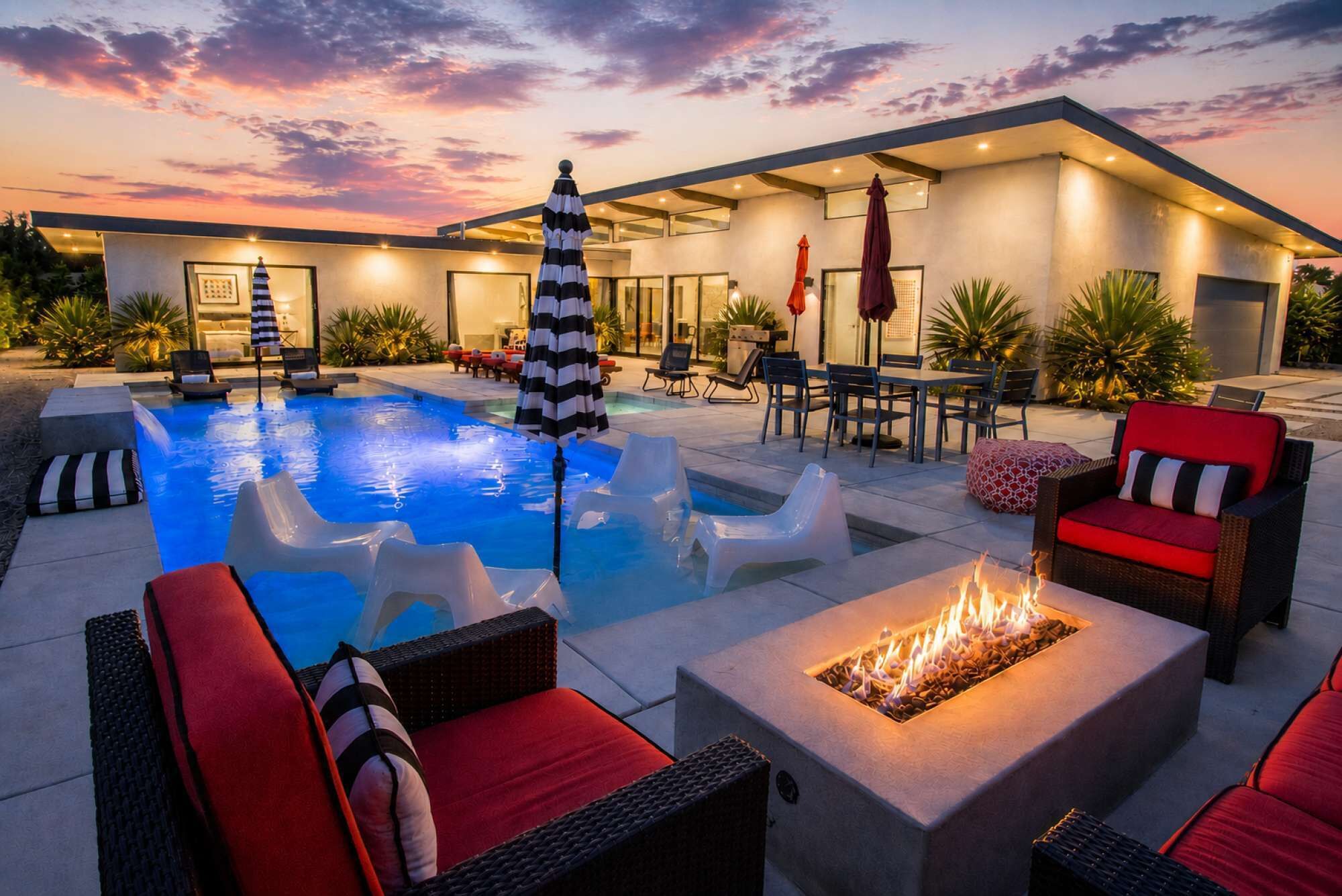 A modern outdoor pool area with lounge chairs, a fire pit, and a sunset view.