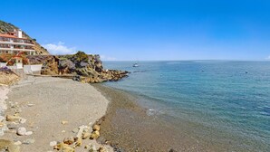 Edifici costaner amb un balcó corbat davant d'un litoral rocos i un mar tranquil.