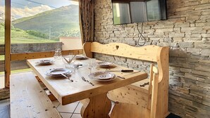 Une table de salle à manger en bois, mise avec des assiettes, des verres et des couverts, un banc en bois et un mur en pierre sur lequel est fixé un téléviseur.