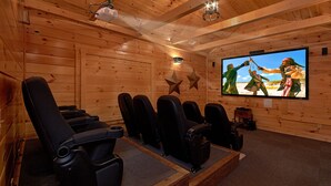 A wooden-paneled room with a large flat-screen TV mounted on the wall, multiple black reclining chairs, and a projector on the ceiling.