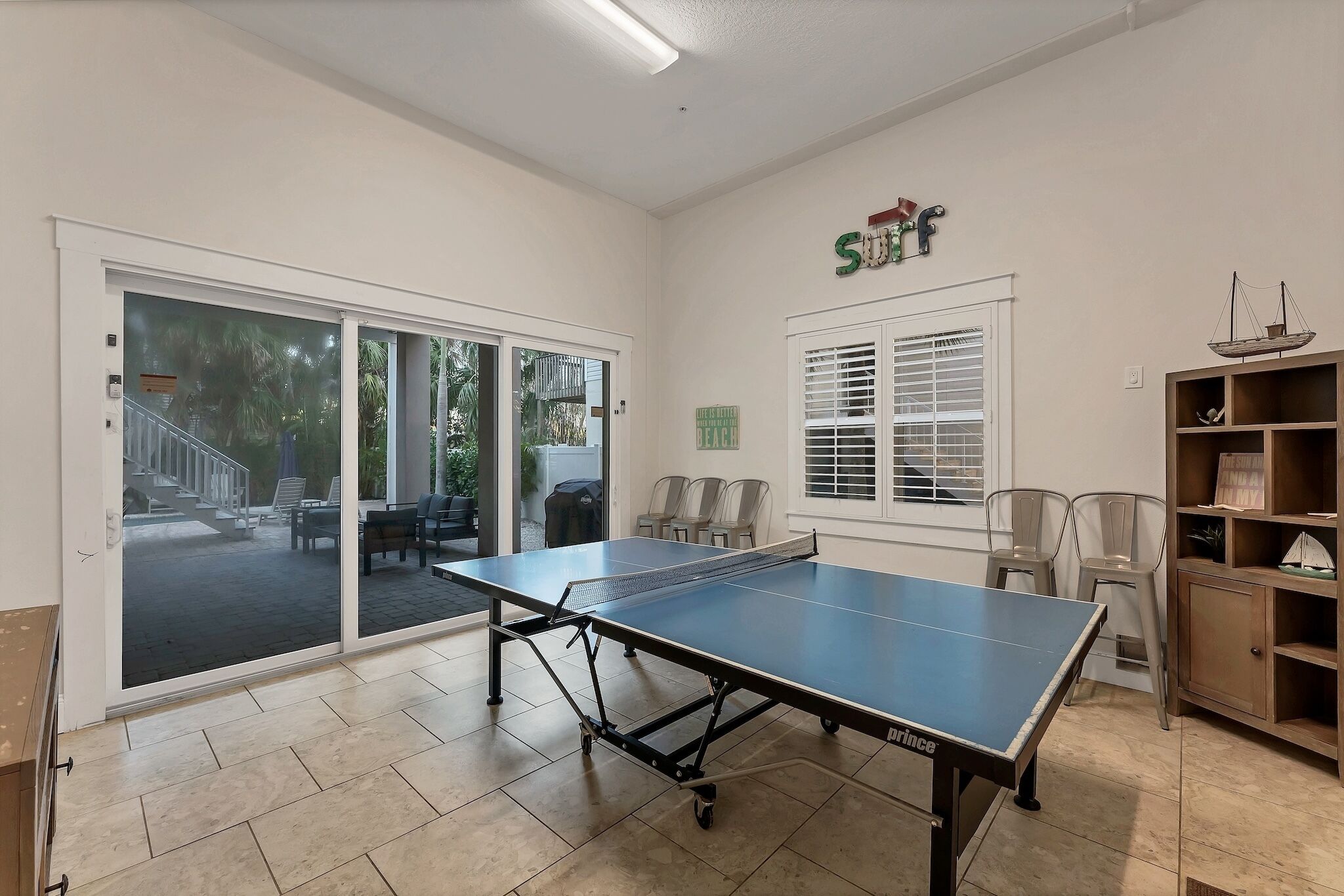 A ping pong table in a room with a sliding glass door leading to an outdoor area with chairs.