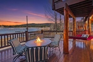 A wooden deck with outdoor seating, a fire pit, and a view of a body of water at dusk.
