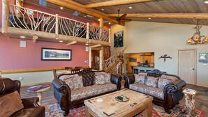 A cozy living room with a high wooden ceiling, a staircase with a rustic design, and a dining area visible through an open door. - Destro's Lakefront by Destination Big Bear : Luxury Lakefront! HOT TUB! PET FRIENDLY 🐾! Pool Table! WiFi! EV Charger! (Big Bear Lake)
