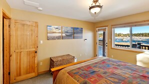 A bedroom with a large bed, a wooden door, a view of the marina through a window, and two wall-mounted paintings. - Destro's Lakefront by Destination Big Bear : Luxury Lakefront! HOT TUB! PET FRIENDLY 🐾! Pool Table! WiFi! EV Charger! (Big Bear Lake)