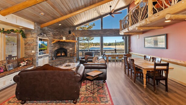 A spacious living room with a stone fireplace, leather sofa, and a dining area with wooden furniture. - Destro's Lakefront by Destination Big Bear : Luxury Lakefront! HOT TUB! PET FRIENDLY 🐾! Pool Table! WiFi! EV Charger! (Big Bear Lake)