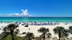 Ein Strand mit Reihen von Sonnenschirmen und Liegestühlen, Palmen und einem strahlend blauen Himmel.