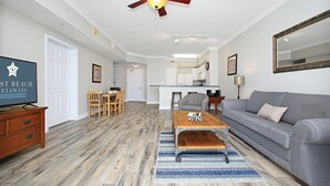A living room with a grey sofa, wooden coffee table, and a dining area with a wooden table and chairs. A ceiling fan and recessed lighting are visible.