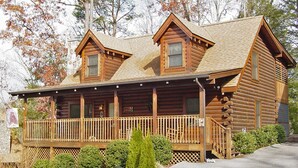 A log cabin with a porch, a chimney, and a stone wall adjacent to it. - ER 246 – The Getaway  Great Location – Close to town! (Pigeon Forge)