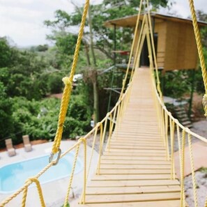 Un puente colgante de madera con cuerdas amarillas, que lleva a una construcción de madera.