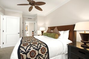 Ein Hotelzimmer mit Bett, zwei Nachttischlampen, einem Nachttisch, einem Deckenventilator und einer Tür zu einem weiteren Zimmer.