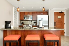 Une cuisine dotée de meubles en bois, d’appareils électroménagers en acier inoxydable, d’un comptoir en granit et de trois tabourets de bar en osier avec des coussins orange.