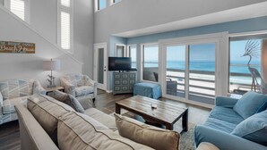 A spacious living room with a large sofa, a coffee table, and a TV. There are multiple windows offering a view of the beach. - Seawinds 1 Beach Front Townhome (Miramar)