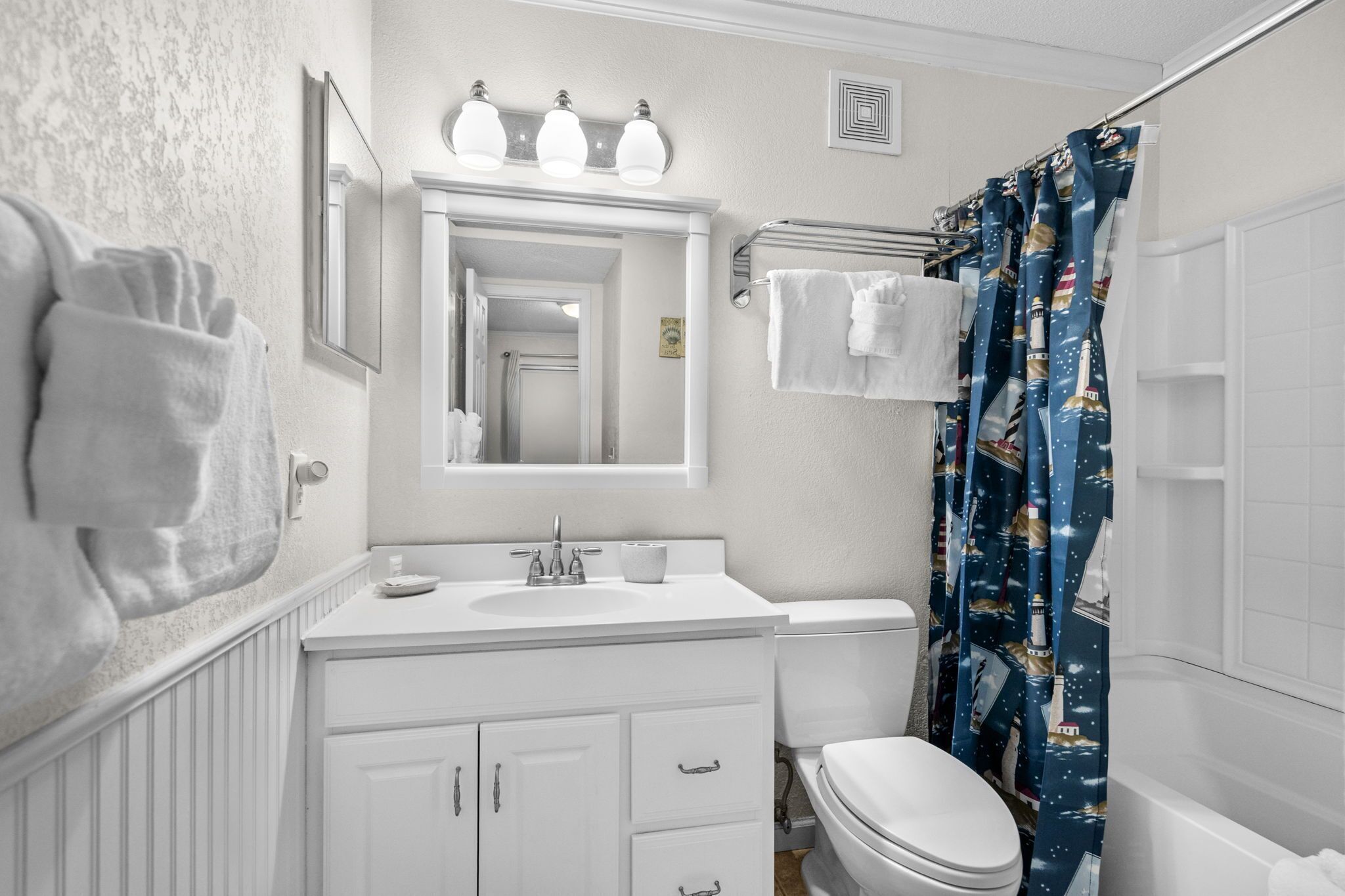 A bathroom with a white vanity, a toilet, a mirror, and a shower curtain with a lighthouse pattern.