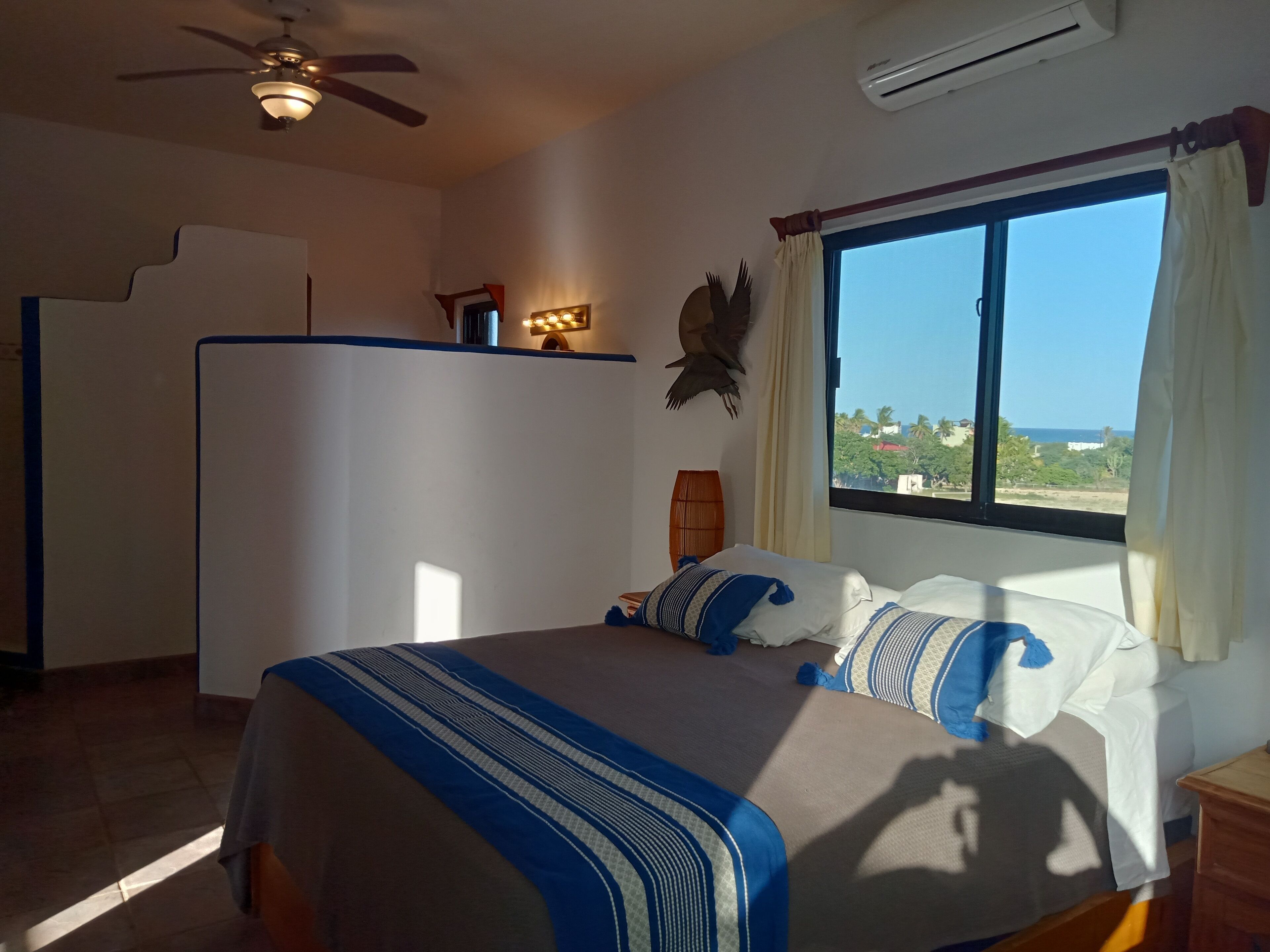 Un dormitorio con cama, ventilador de techo, aire acondicionado, ventana con cortinas y vistas al exterior.
