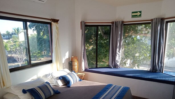 A bedroom with a bed, a window with curtains, a view of greenery outside, and a sign indicating an evacuation route. - Hacienda de Palmas: Room #2 Cupola with one queen size bed (La Ribera)