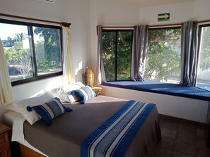 A bedroom with a bed, a window with curtains, a view of greenery outside, and a sign indicating an evacuation route. - Hacienda de Palmas: Room #2 Cupola with one queen size bed (La Ribera)