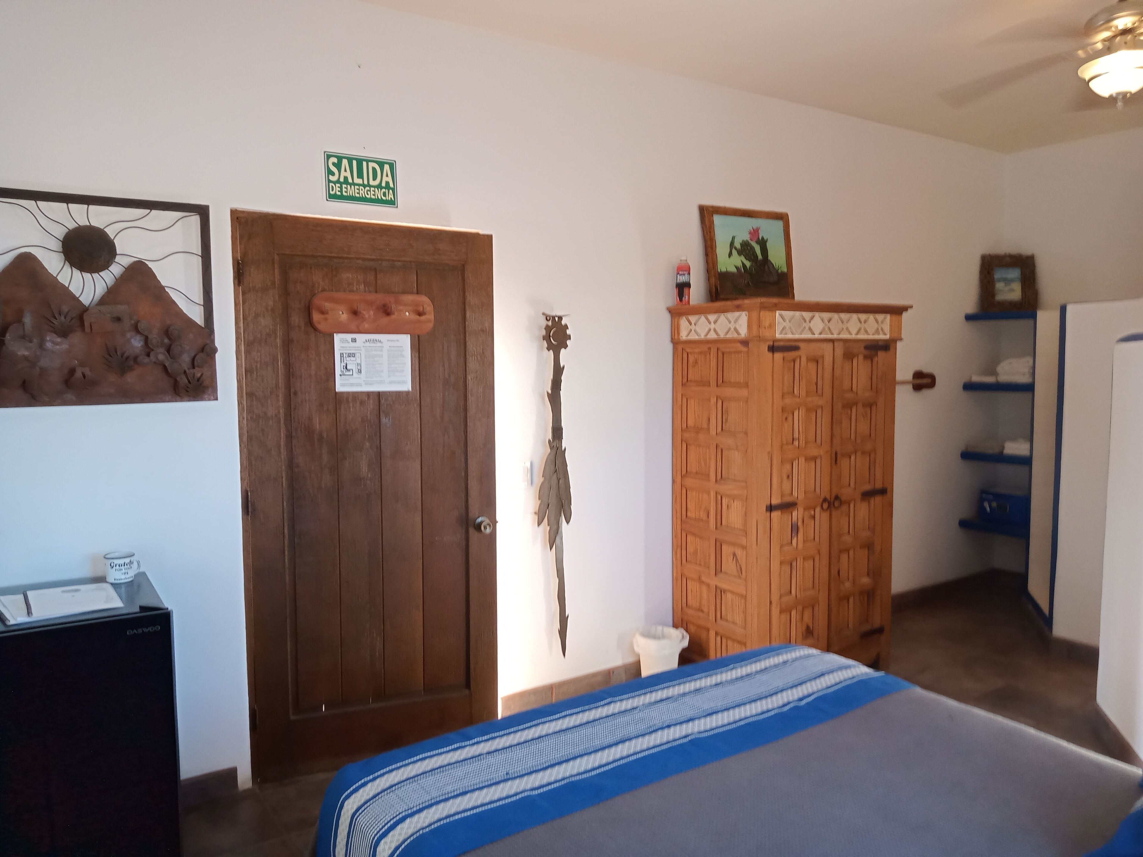 A room with a bed, a wooden wardrobe, a door with an emergency exit sign, and a wall-mounted decoration.