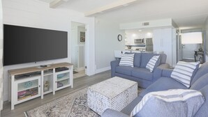 Una sala de estar moderna con un televisor de pantalla plana, un conjunto de sofá gris, una mesa de centro y una cocina al fondo.
