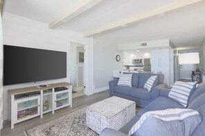 Un salon moderne avec un téléviseur à écran plat, un ensemble de canapés gris, une table basse et une cuisine en arrière-plan.