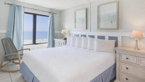 Habitación de hotel con una cama grande, una silla, una mesita de noche, un aparador y vistas a la playa.