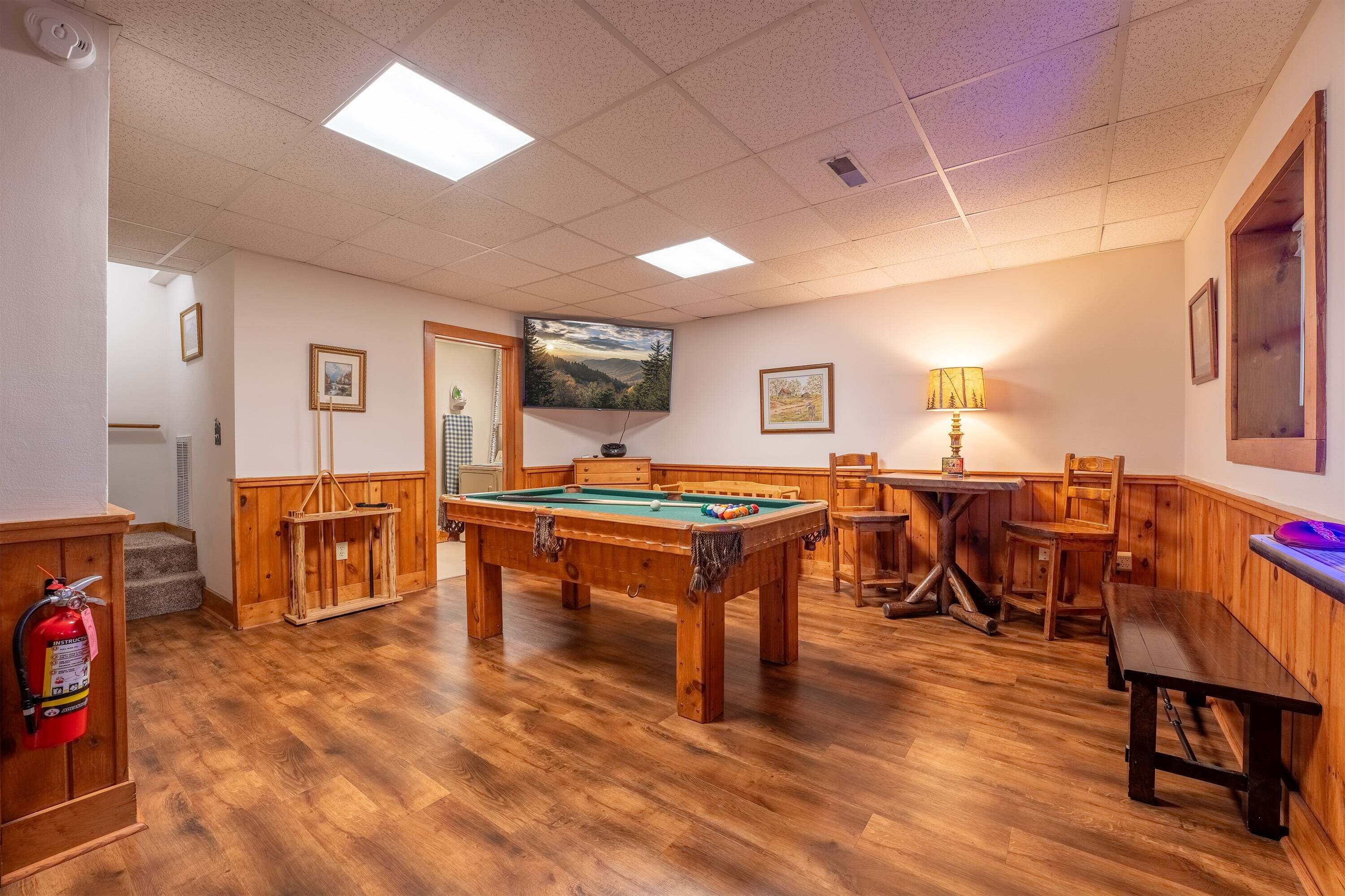 Une table de billard dans une pièce dont le sol est recouvert de parquet, avec un extincteur, un téléviseur et des tableaux encadrés accrochés aux murs.