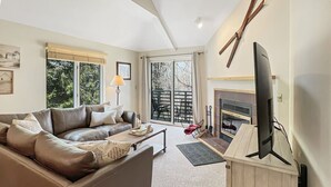 A living room with a sofa, a coffee table, a TV on a wooden stand, and a fireplace. - Woods  Resort Village 10 Cozy Killington Escape –1BR + Loft, Fireplace, Pool & Hot Tub Access (Killington)